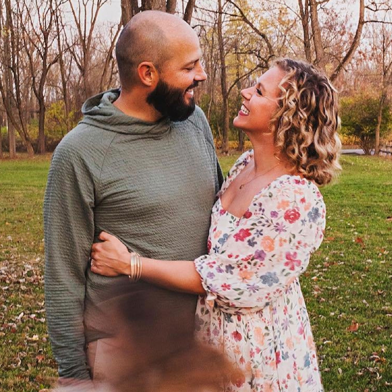 A couple stand together in an autumn park while their kids run around.