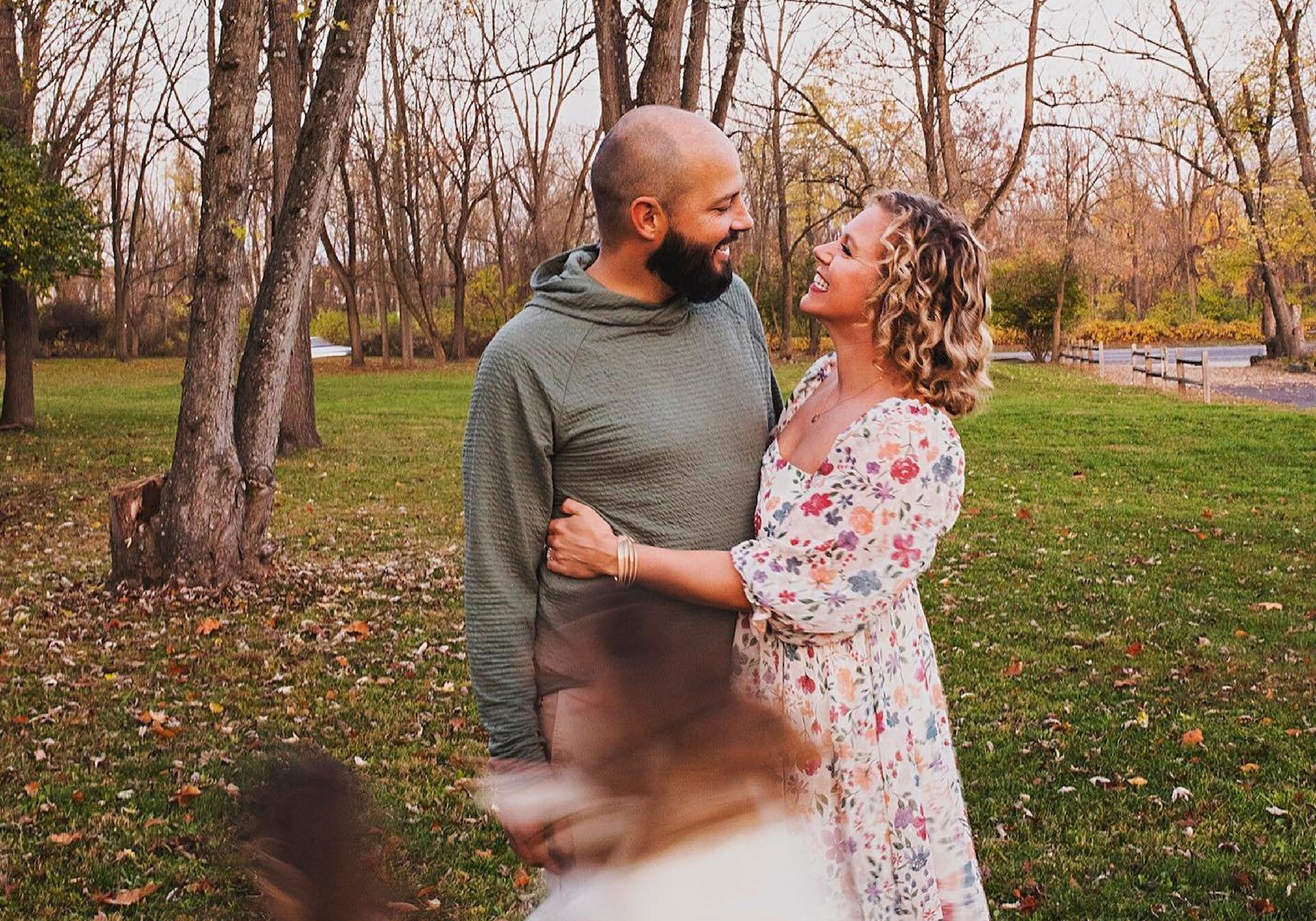 A couple stand together in an autumn park while their kids run around.