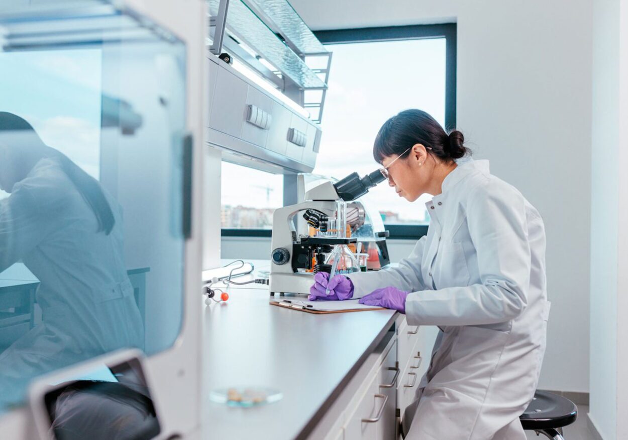 A scientist in a lab looking through a microscope and writing on a clipboard.