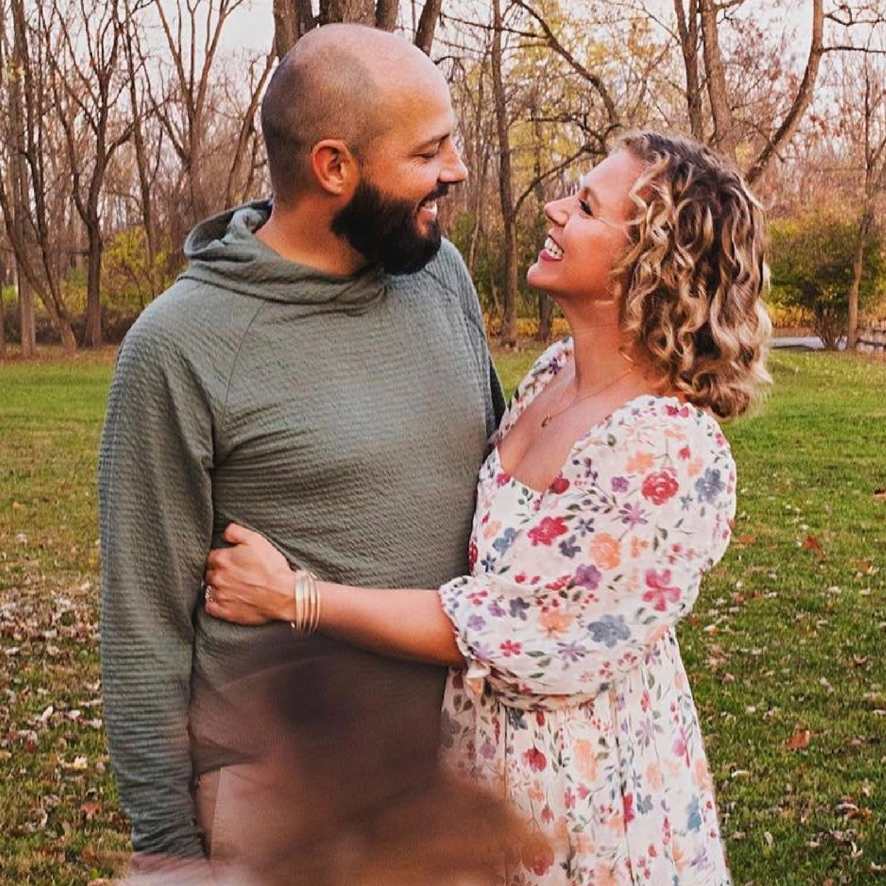 A couple stand together in an autumn park while their kids run around.