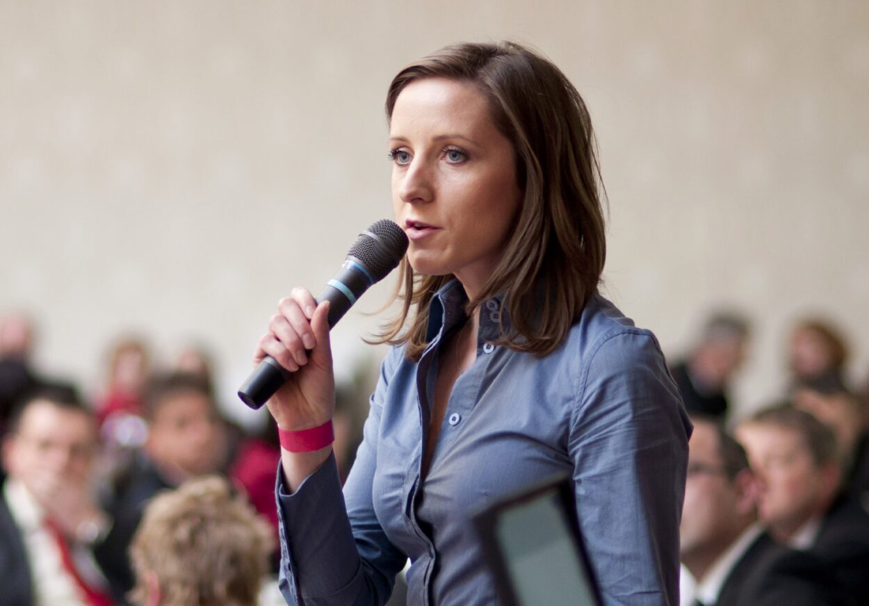 woman with microphone presenting to a crowded room.
