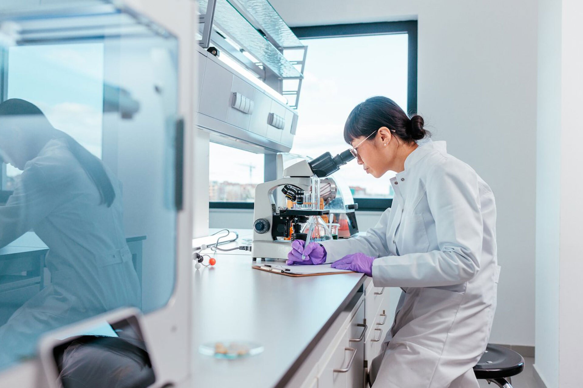 A scientist in a lab looking through a microscope and writing on a clipboard.